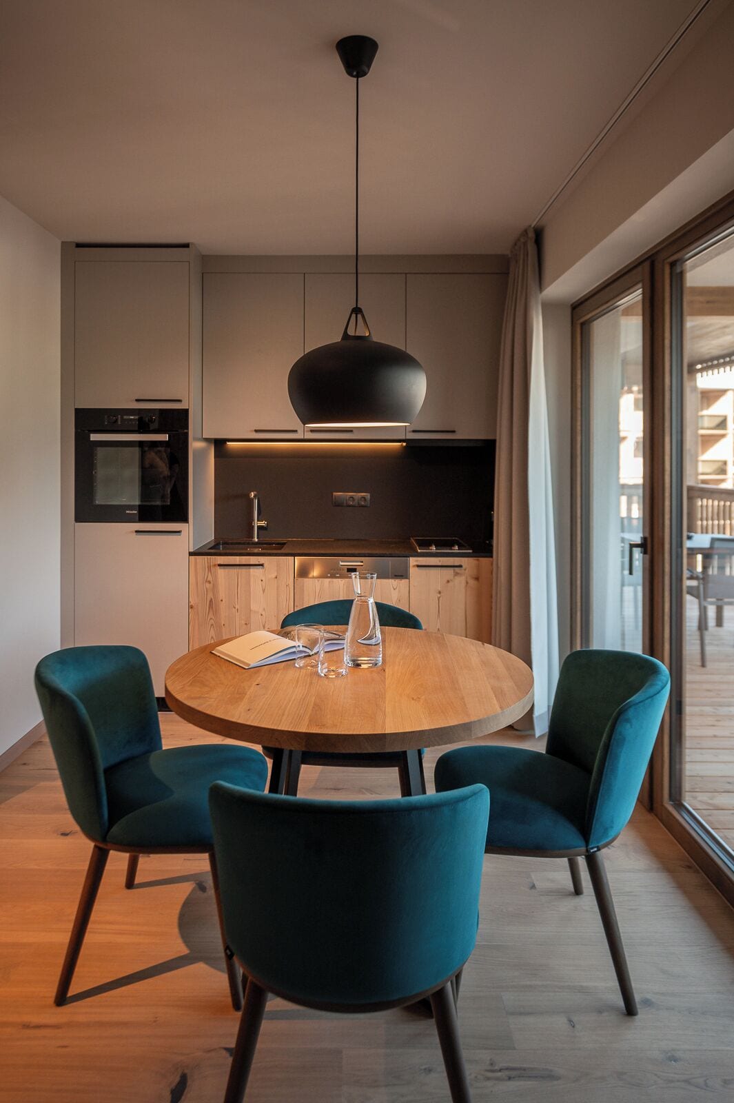 A modern kitchen with a round wooden table, teal chairs, and a pendant light.