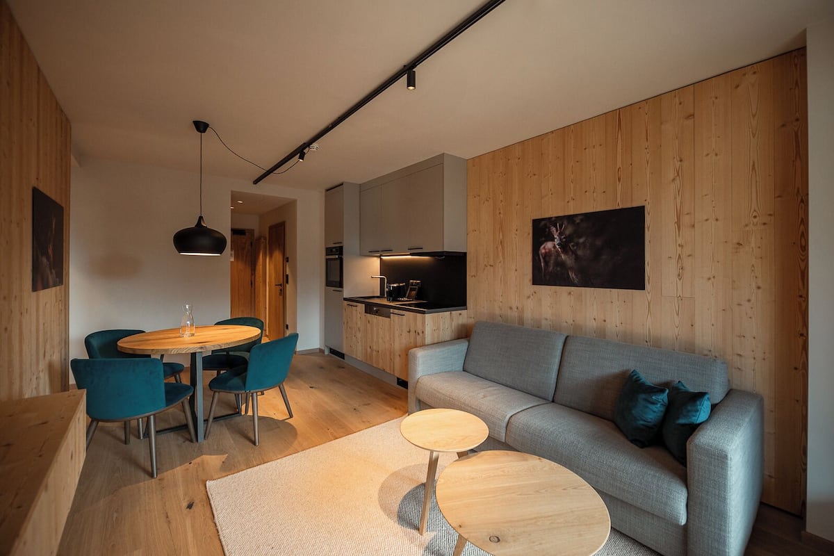 Una sala moderna con un sofá gris, sillas color turquesa, una mesa de comedor de madera y una zona de cocina con televisión.