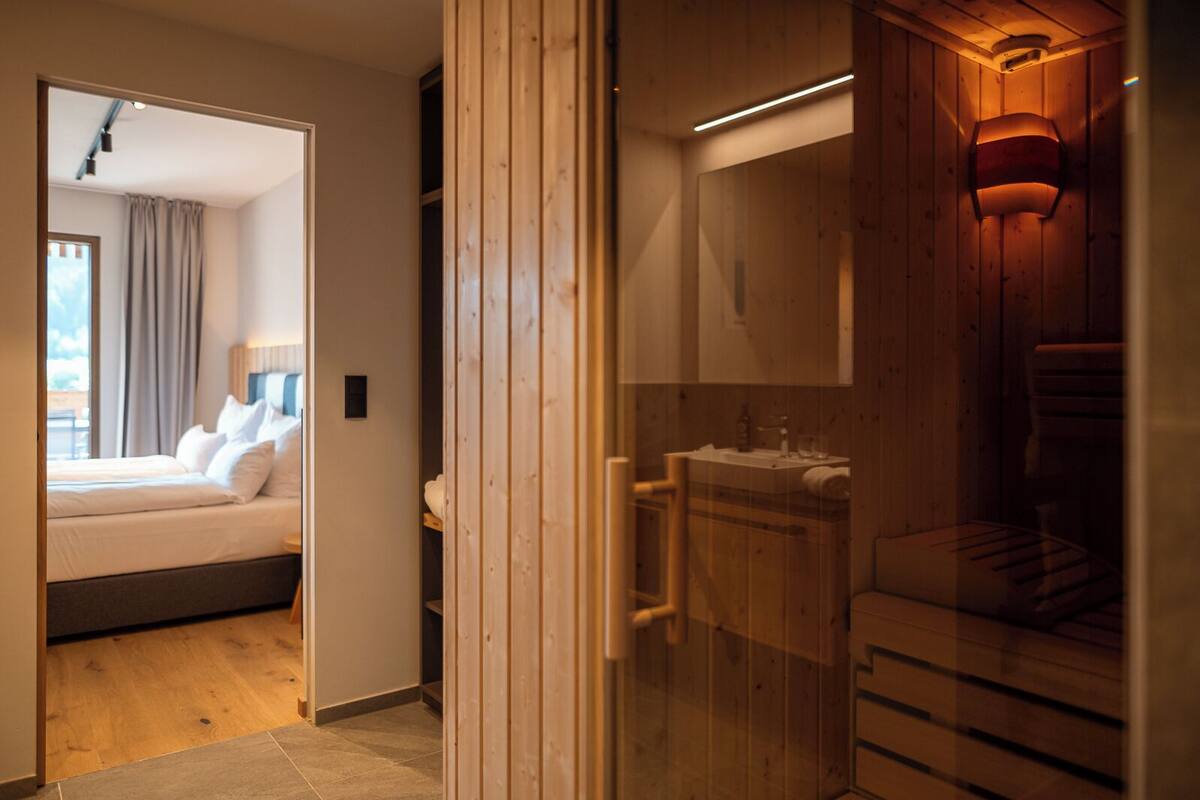 Habitación de hotel moderna con detalles de madera, una cama con ropa blanca y un baño con tocador de madera, además de vistas al exterior.