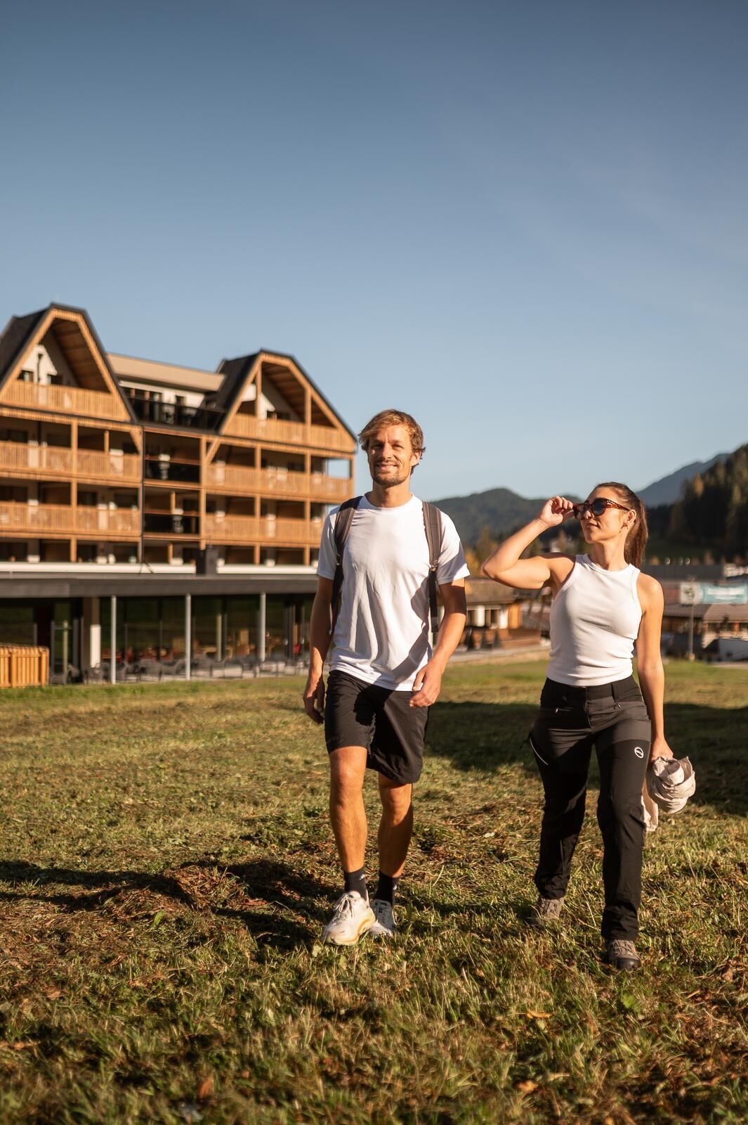 Un uomo e una donna camminano su un prato erboso, con un edificio in legno sullo sfondo.