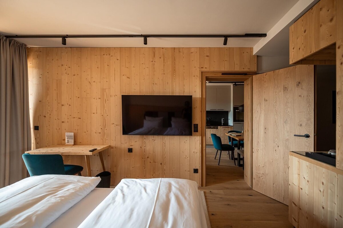 A modern hotel room with wooden panel walls, a flat-screen TV mounted above a bed, a desk with a chair, and a view into the kitchen area.