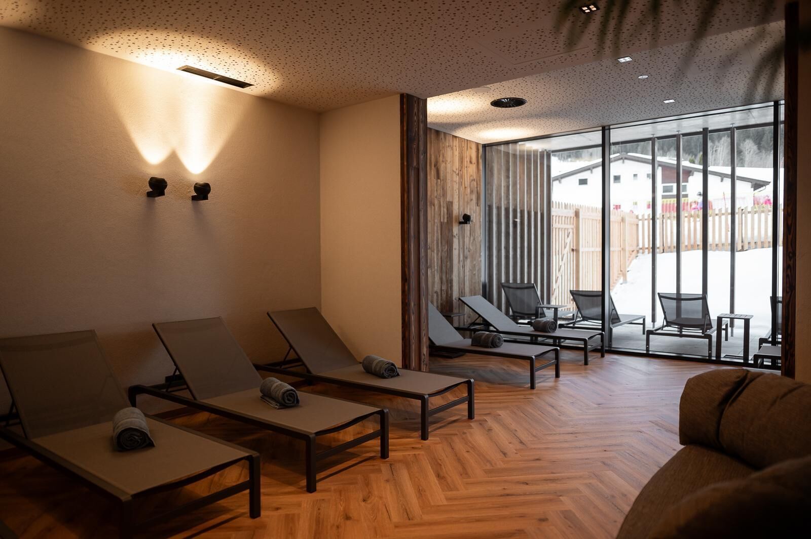 A modern lounge area with wooden flooring, sun loungers, and a glass door leading to an outdoor seating area.