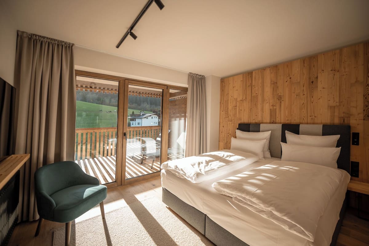 Un dormitorio con una cama grande, un escritorio y una silla. Hay un ventanal corredero que da a un balcón con vistas a un paisaje verde y un edificio.