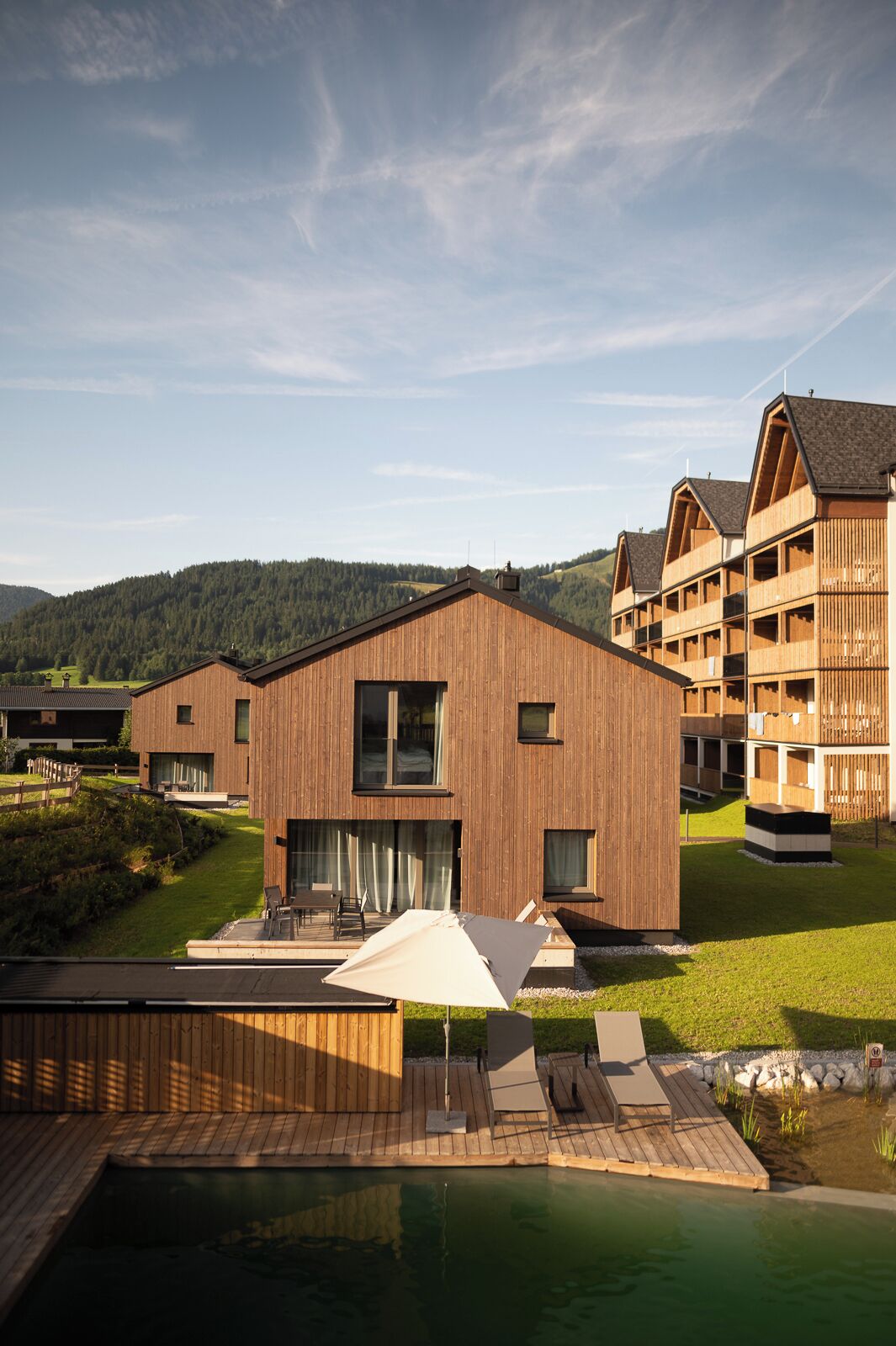 A modern wooden building with a pool area, surrounded by a lush green landscape and hills in the background.