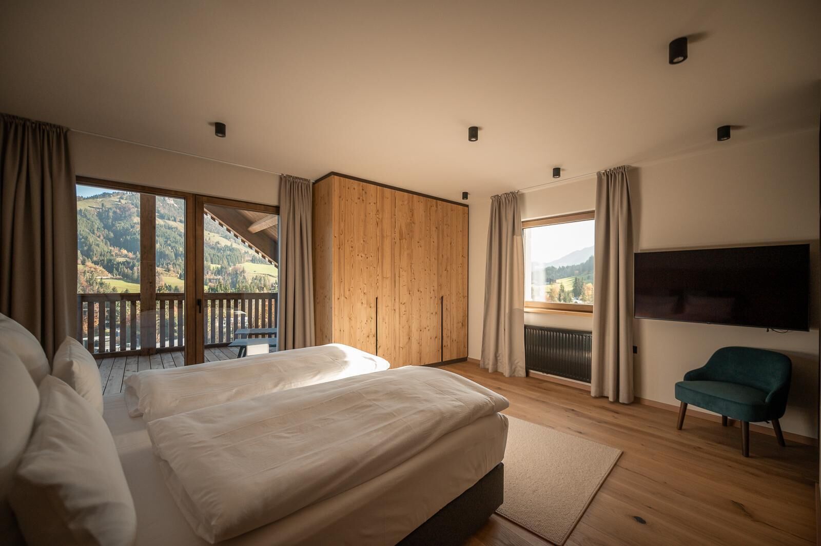 A bedroom with a large bed, a TV, a chair, and a balcony with a view of the mountains.