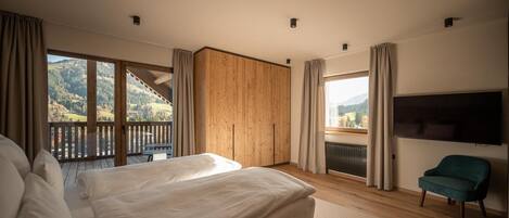 A bedroom with a large bed, a TV, a chair, and a balcony with a view of the mountains.