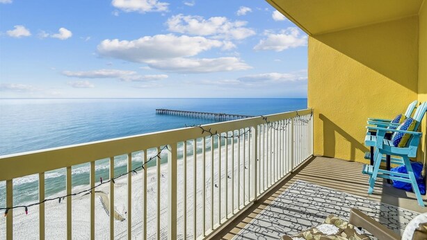 A balcony with a view of the ocean and a pier. There are blue chairs and a patterned rug on the balcony floor.