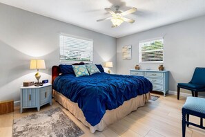 Un dormitorio con cama, mesitas de noche, una silla, ventilador de techo y ventanas con persianas.