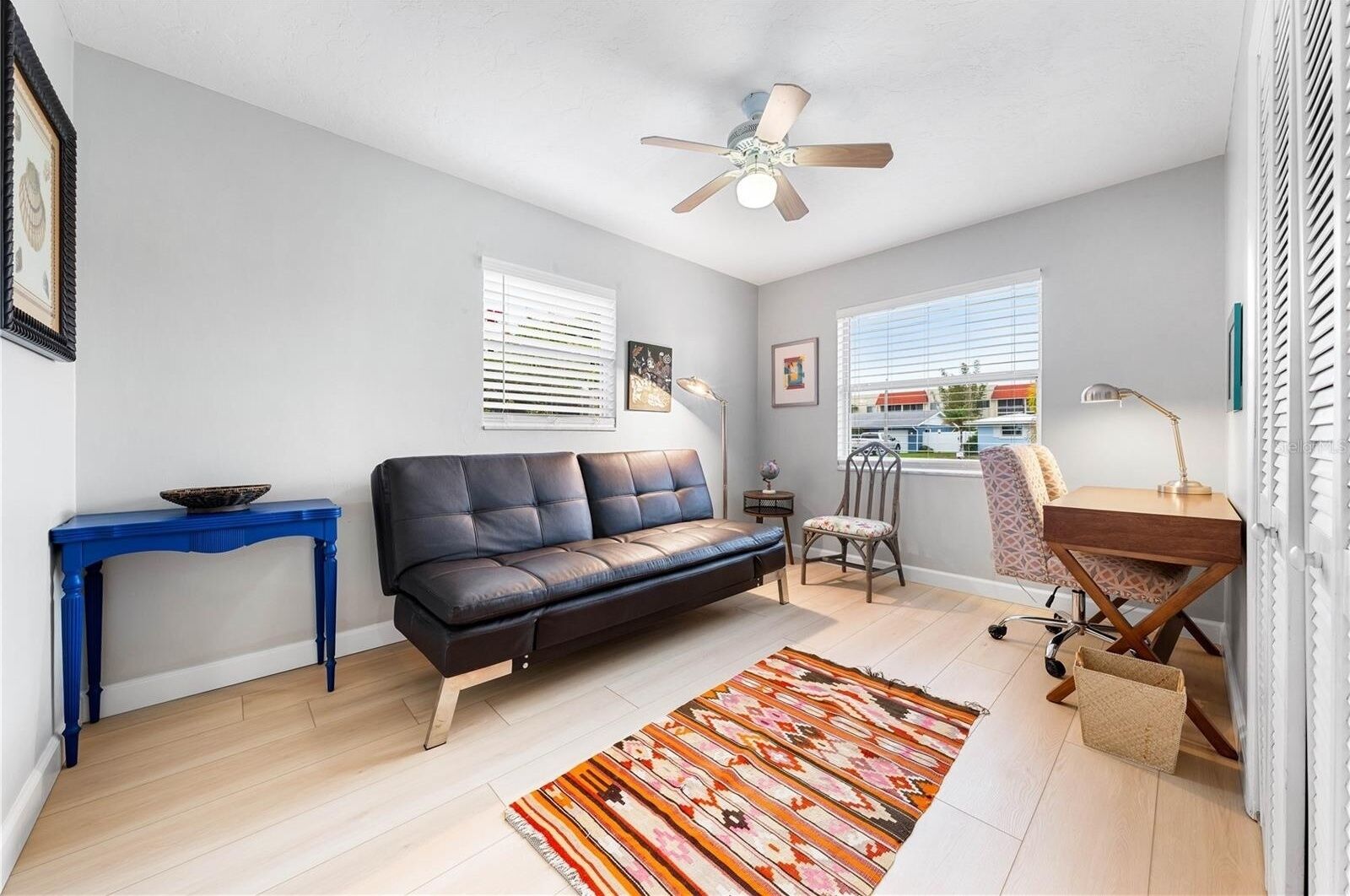 A modern living room with a sofa, a desk, a chair, a blue console table, a patterned rug, and a ceiling fan.