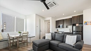Un salon moderne comprenant un canapé d’angle, une table à manger avec des chaises et un coin cuisine doté de meubles et d’appareils électroménagers aux couleurs sombres.