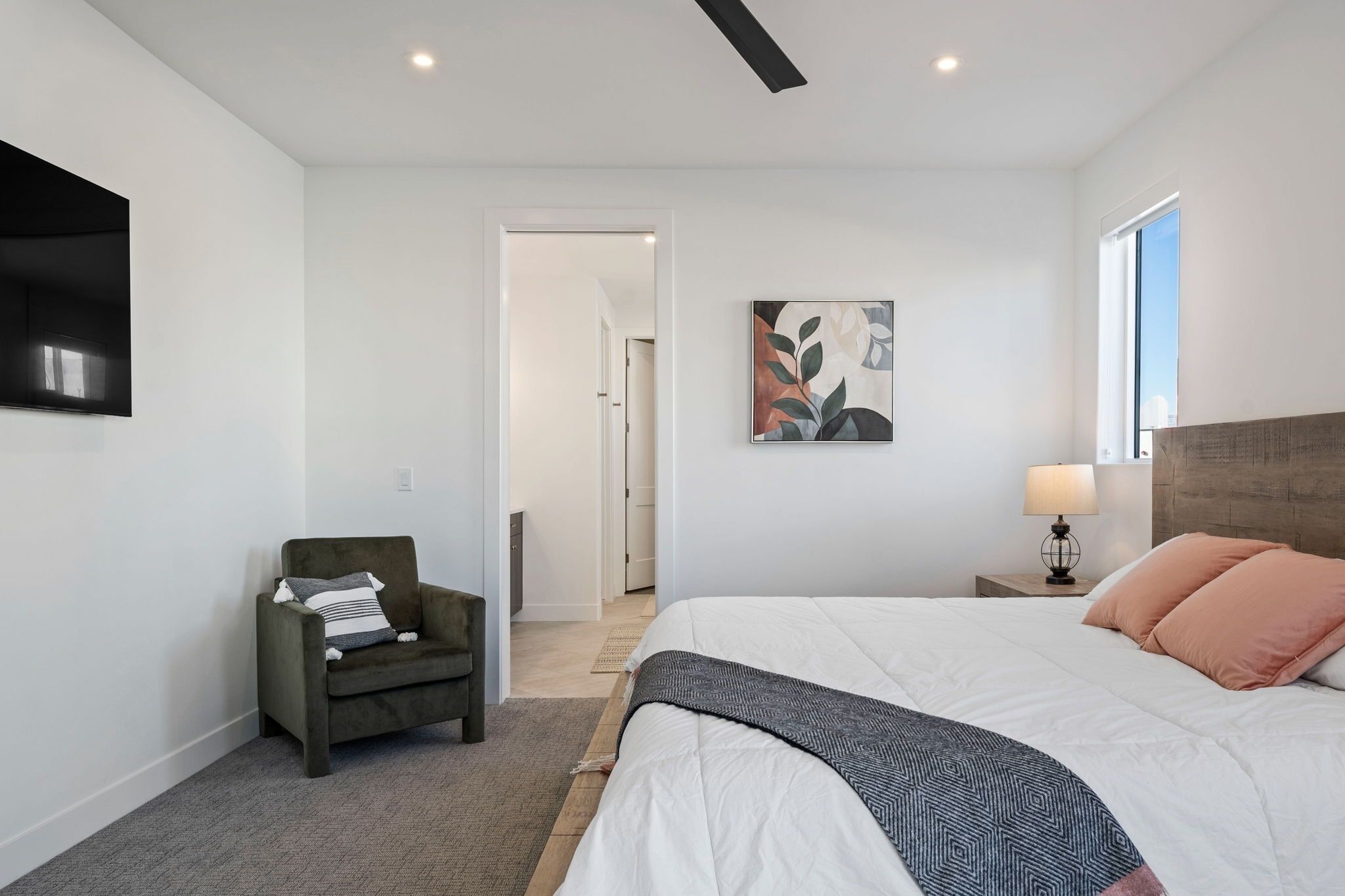 A bedroom with a bed, a chair, a bedside table with a lamp, a painting on the wall, and a door leading to another room.
