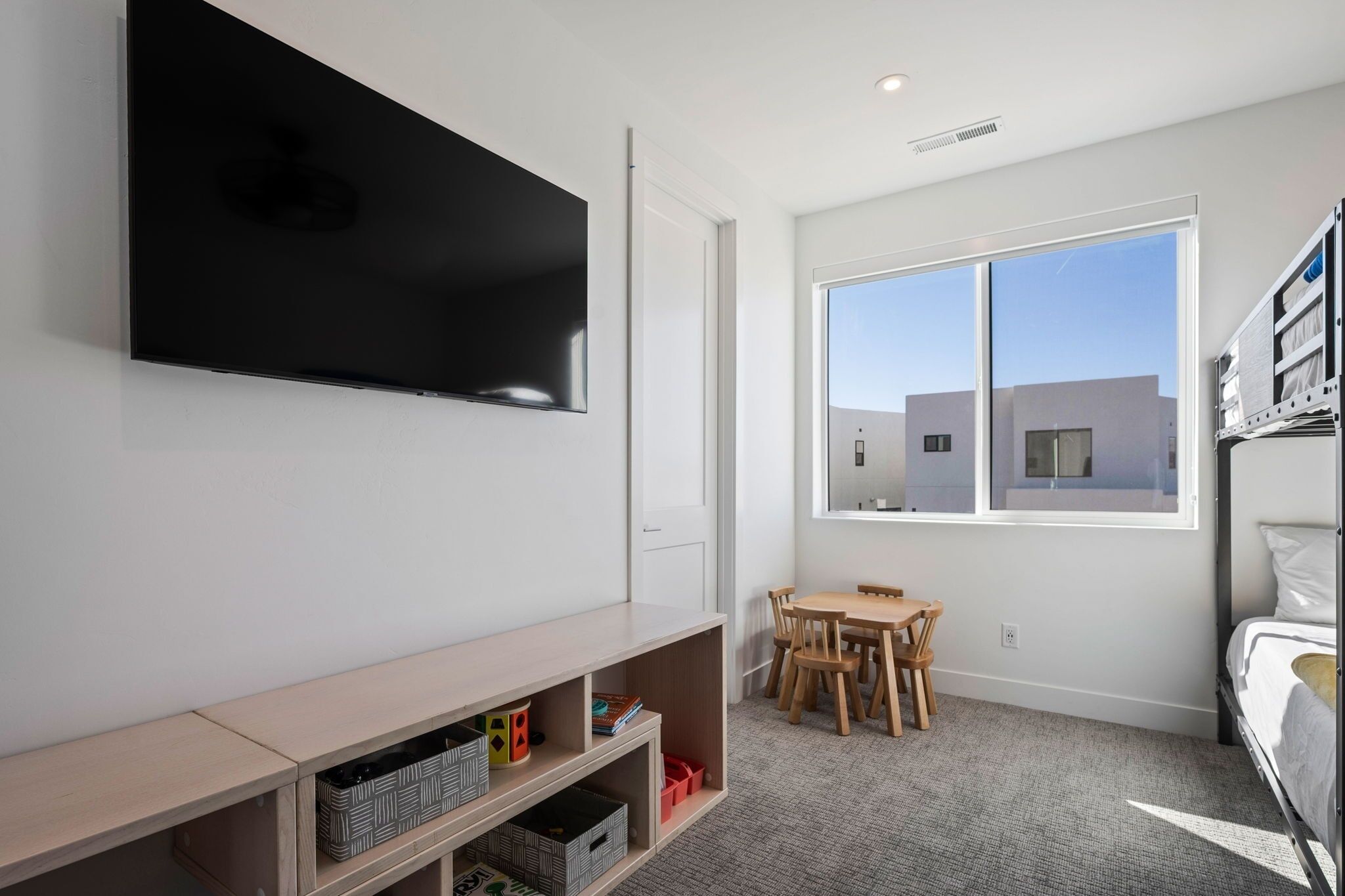 A room with a TV mounted on the wall, a wooden table with chairs, a bunk bed, and a large window with a view of buildings.