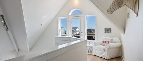 A white interior with a sloped ceiling, a white sofa with a 'PEACE' pillow, and a view of the outside through large windows.