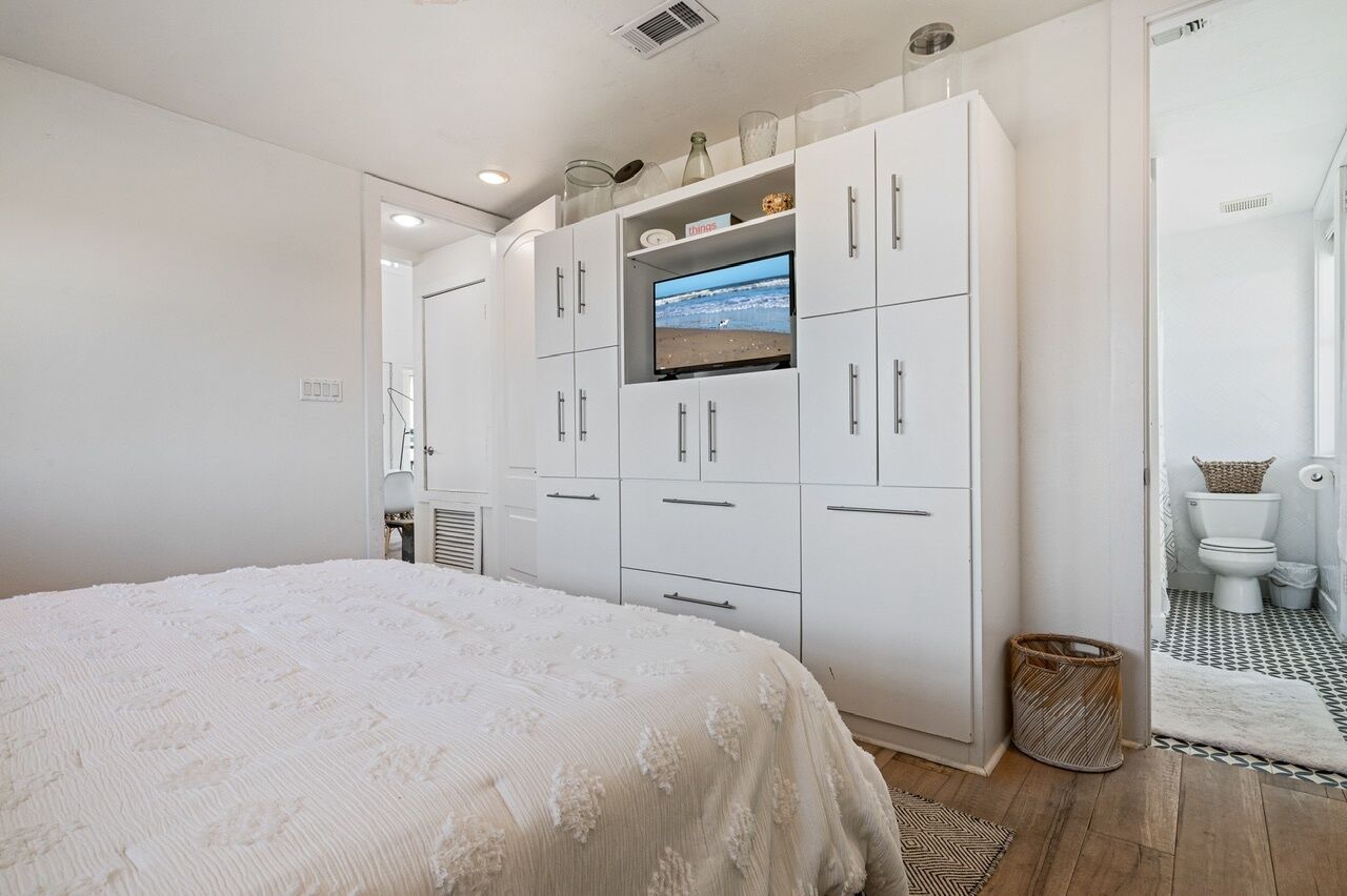 Ein Schlafzimmer mit einem großen Bett, einem an eine weiße Garderobe montierten Fernseher und einem durch eine offene Tür einsehbaren Badezimmer.