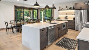 Une cuisine moderne dotée d’un grand îlot central, de meubles de couleur sombre, d’appareils électroménagers en acier inoxydable, de luminaires suspendus et offrant une vue sur la verdure grâce à de grandes fenêtres.