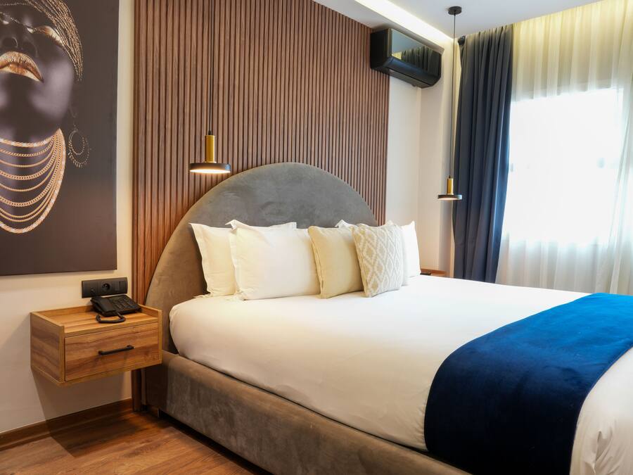 Una habitación de hotel moderna con una cama grande, una mesita de noche de madera con un teléfono, un televisor montado en la pared y un amplio ventanal con cortinas.