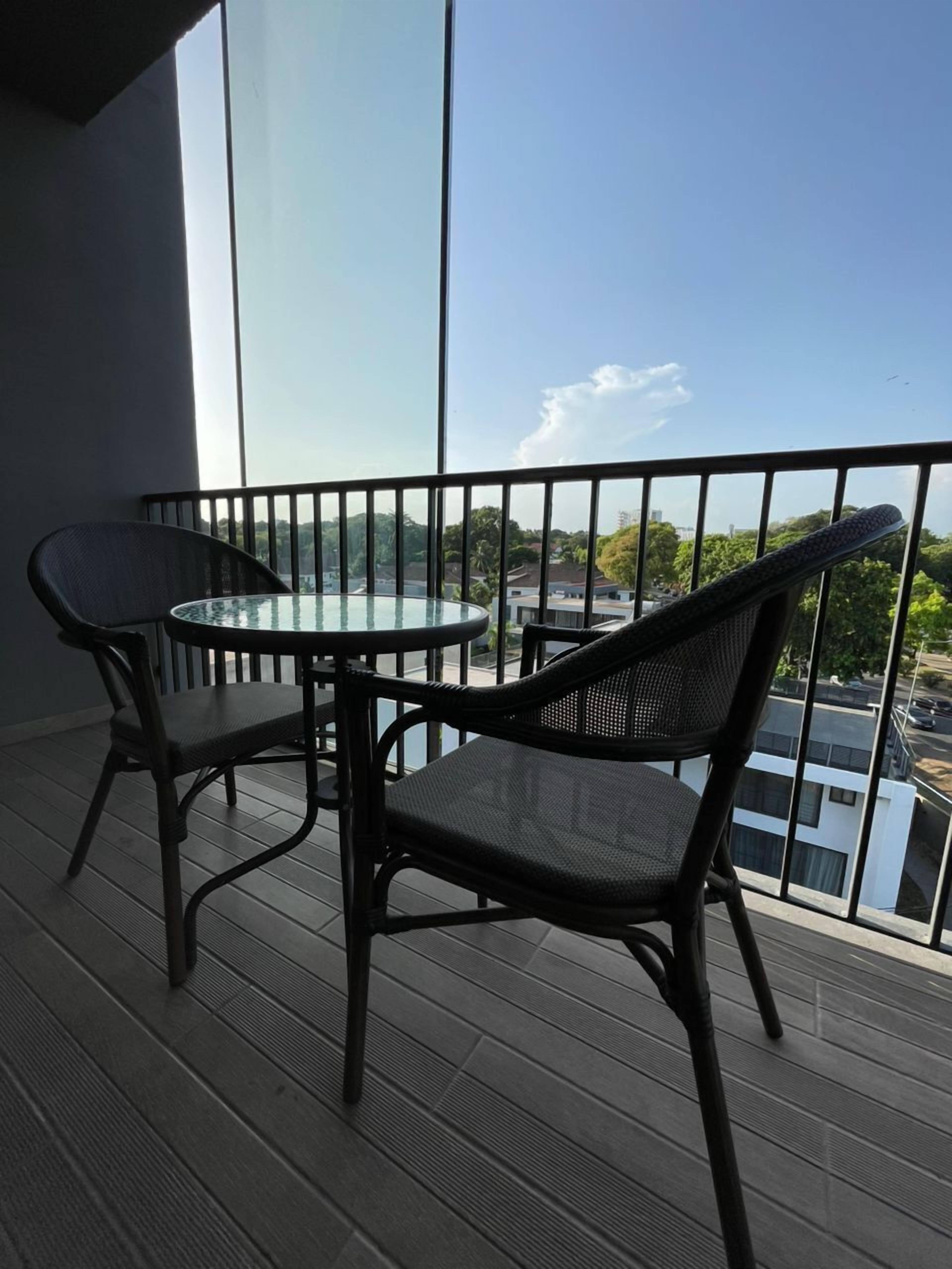 A balcony with a glass table and two wicker chairs, overlooking a cityscape.