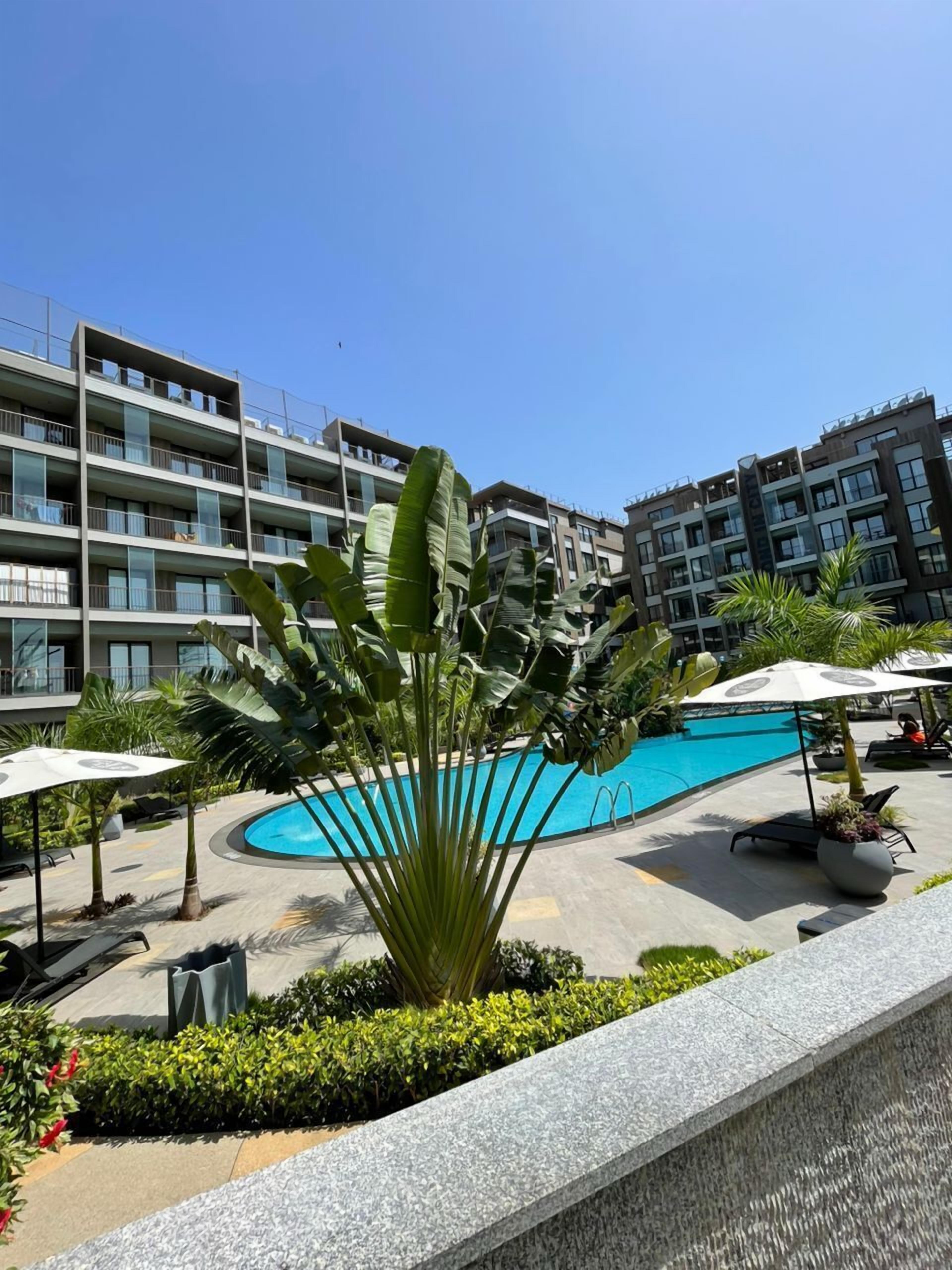 A swimming pool surrounded by lounge chairs and umbrellas, with multi-story buildings in the background.