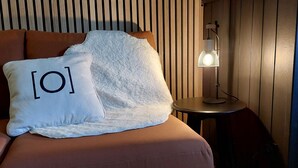 Ein Bett mit brauner Kopfteil, ein weißes Kopfkissen mit schwarzem Text und ein kleiner Tisch mit einer Lampe daneben.