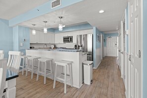 Una cocina moderna con muebles blancos, una isla central con taburetes de bar, electrodomésticos de acero inoxidable y una pared de acento en tono azul claro.