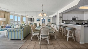 A spacious living area with a large sofa, dining table, and chairs, a kitchen with white cabinets and a bar area. - Oceanfront Crescent Beach 1K 2Q SS | 4th Floor Unit 409 (North Myrtle Beach)