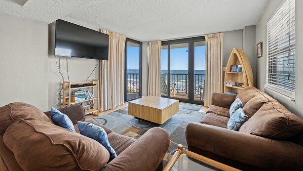 A modern living room with a flat-screen TV, brown sofas, a wooden coffee table, and a bookshelf. Large windows offer a view of the beach. - Oceanfront Crescent Beach 1K, 1Q, 1D, 2Tw, SS (North Myrtle Beach)