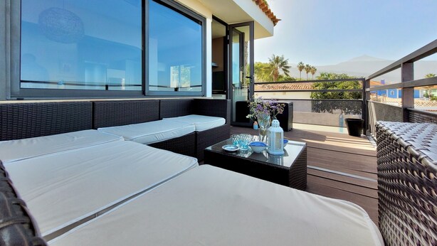 A balcony with wicker furniture, a glass table with decorative items, and a view of palm trees and mountains. - Villa with Privacy and Spectacular Views of Teide, Sea, Orotava Valley (La Orotava)