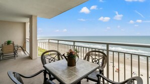 A balcony with a table and chairs, overlooking a beach with the ocean and a clear sky. - Oceanfront Crescent Beach 2K 2T (North Myrtle Beach)