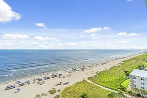 Een strand met parasols en mensen, een gazon en een gebouw met een balkon.