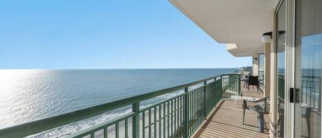 A balcony with a view of the ocean, a wooden deck, and a glass door leading to an outdoor area with chairs.