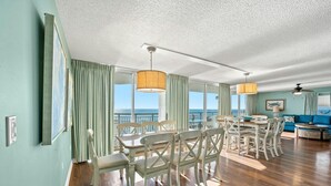 A spacious living area with a dining table and chairs, a blue sofa, and a balcony with a view of the ocean. - 5-bedroom, 4-bathroom oceanfront Penthouse, Sleeps 14 | Windy Hill Dunes - 1803PH (North Myrtle Beach)