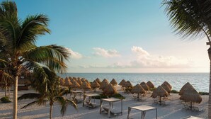 A beach with thatched umbrellas and lounge chairs, a palm tree, and a view of the ocean. - Excellence Coral Playa Mujeres - Adults Only All Inclusive (Punta Sam)
