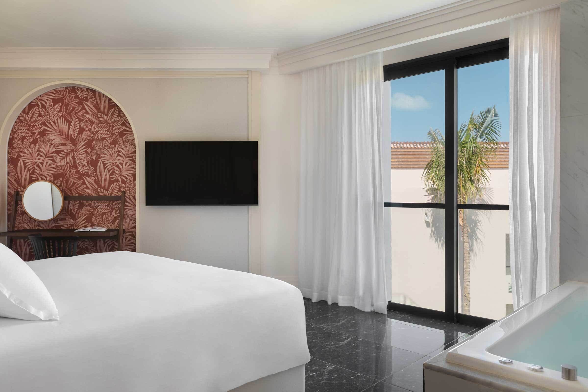 A hotel room with a bed, a TV mounted on the wall, a large window with curtains, and a view of palm trees.