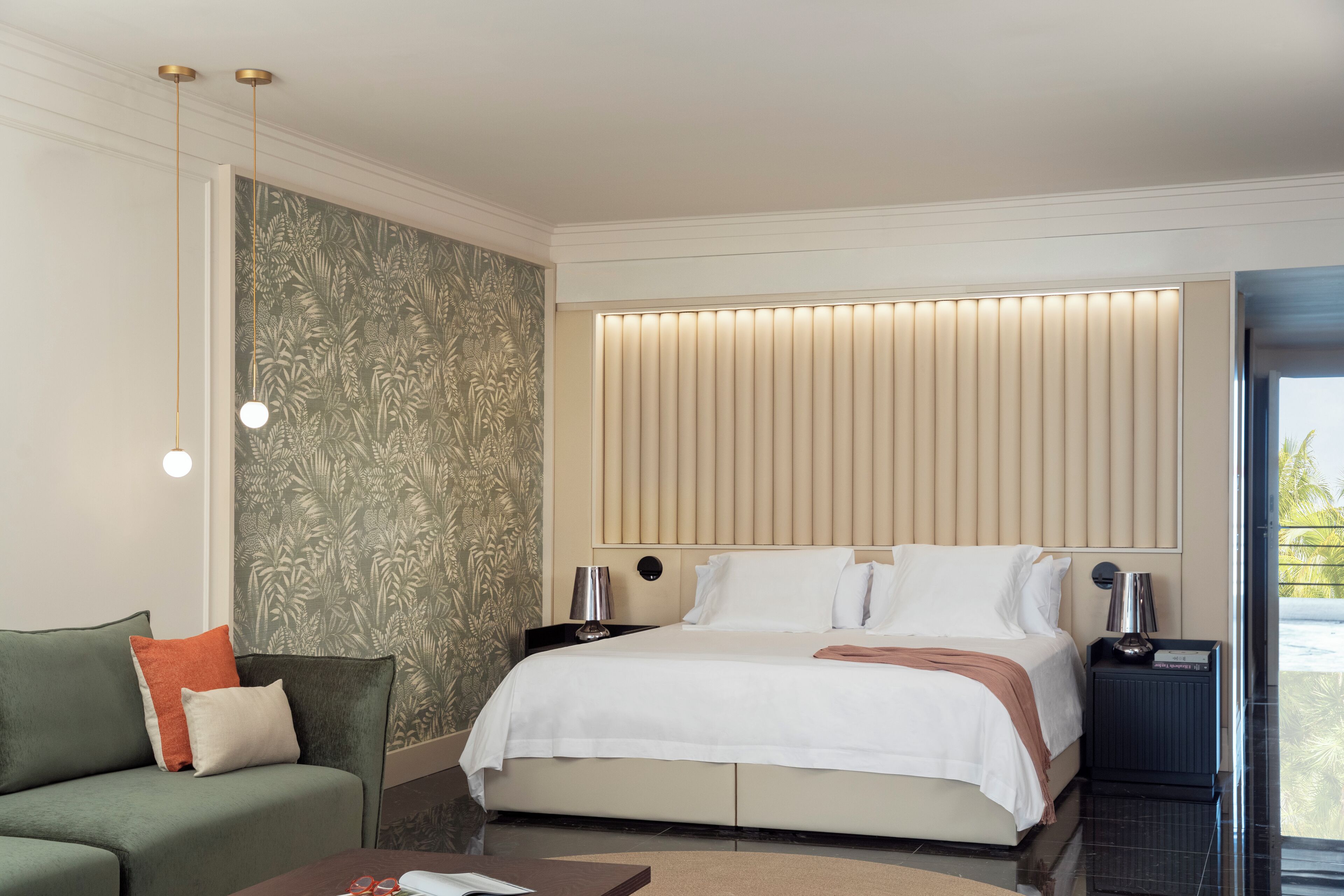 A modern hotel room with a large bed, a sofa, a nightstand with a lamp, and a view of the outdoors.