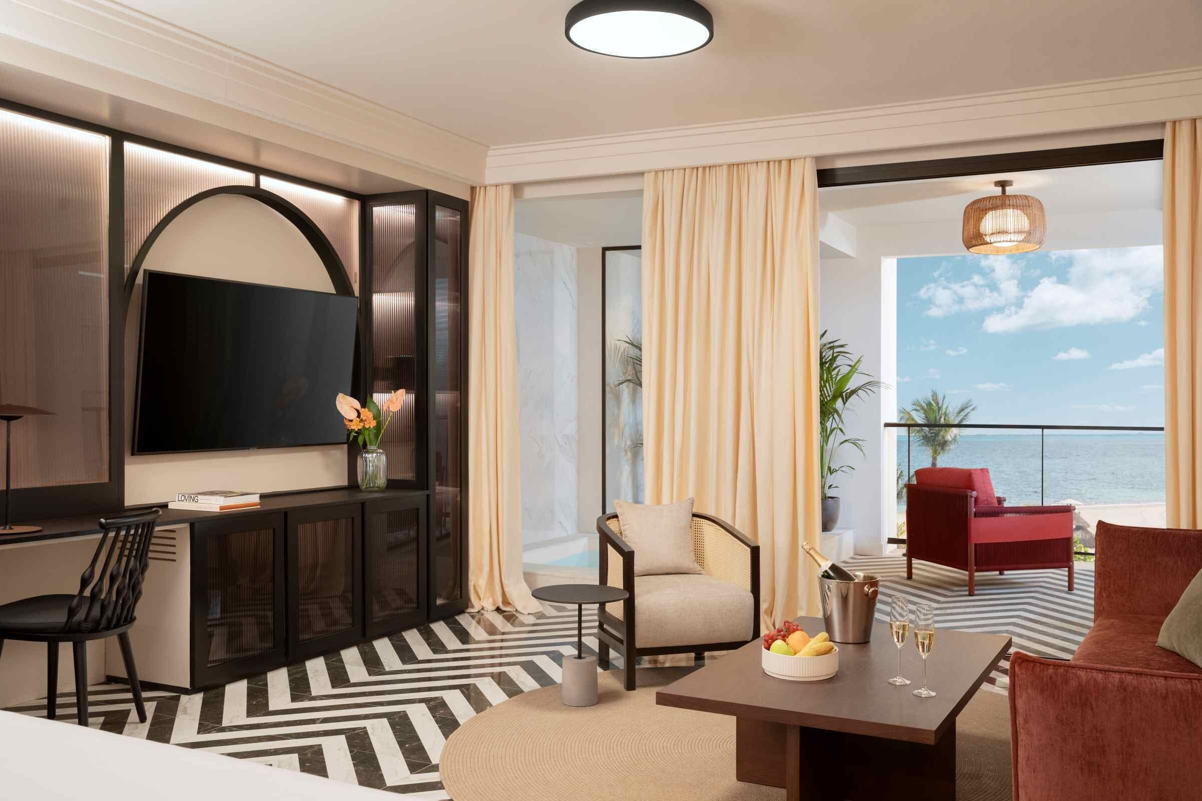 Een moderne hotelkamer met een grote televisie, een comfortabele zithoek, een kleine tafel met een schaal fruit en glazen, en uitzicht op zee.