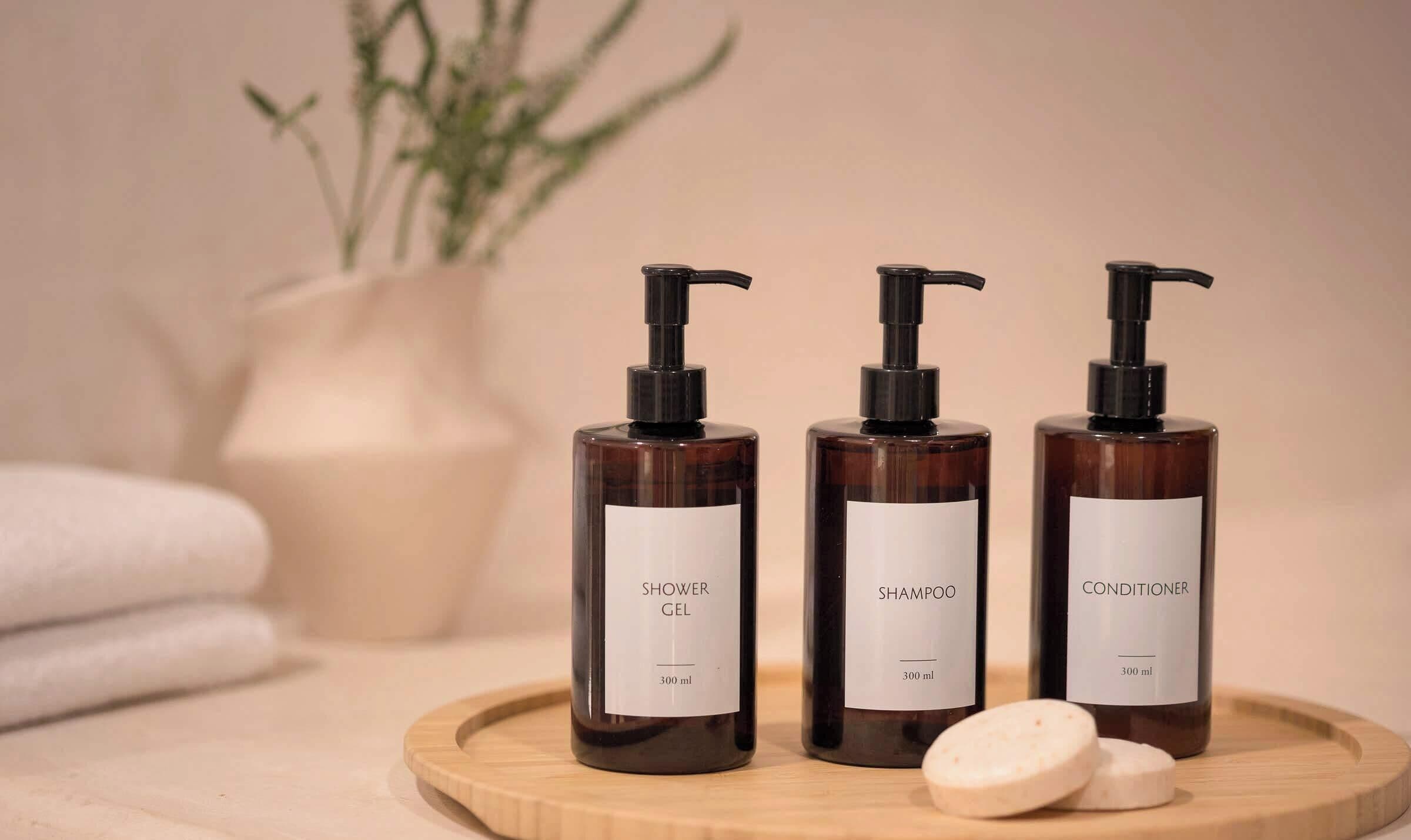 Trois flacons de gel douche, de shampoing et d’après-shampoing, présentés sur un plateau en bois avec deux savons.