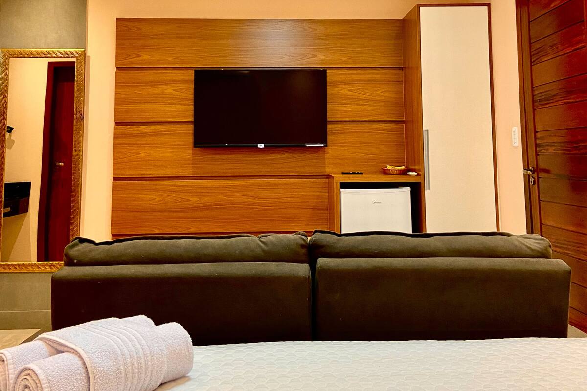 Ein modernes Hotelzimmer mit Flachbildfernseher an einer Wand aus Holzpaneelen, einer Couch, einem kleinen Schreibtisch mit Kühlschrank und einem großen Spiegel.