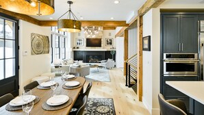 Une cuisine moderne avec des meubles sombres et des appareils en acier inoxydable, un coin repas avec une table en bois pour quatre personnes, et un espace de vie chaleureux avec une cheminée et des sièges confortables.