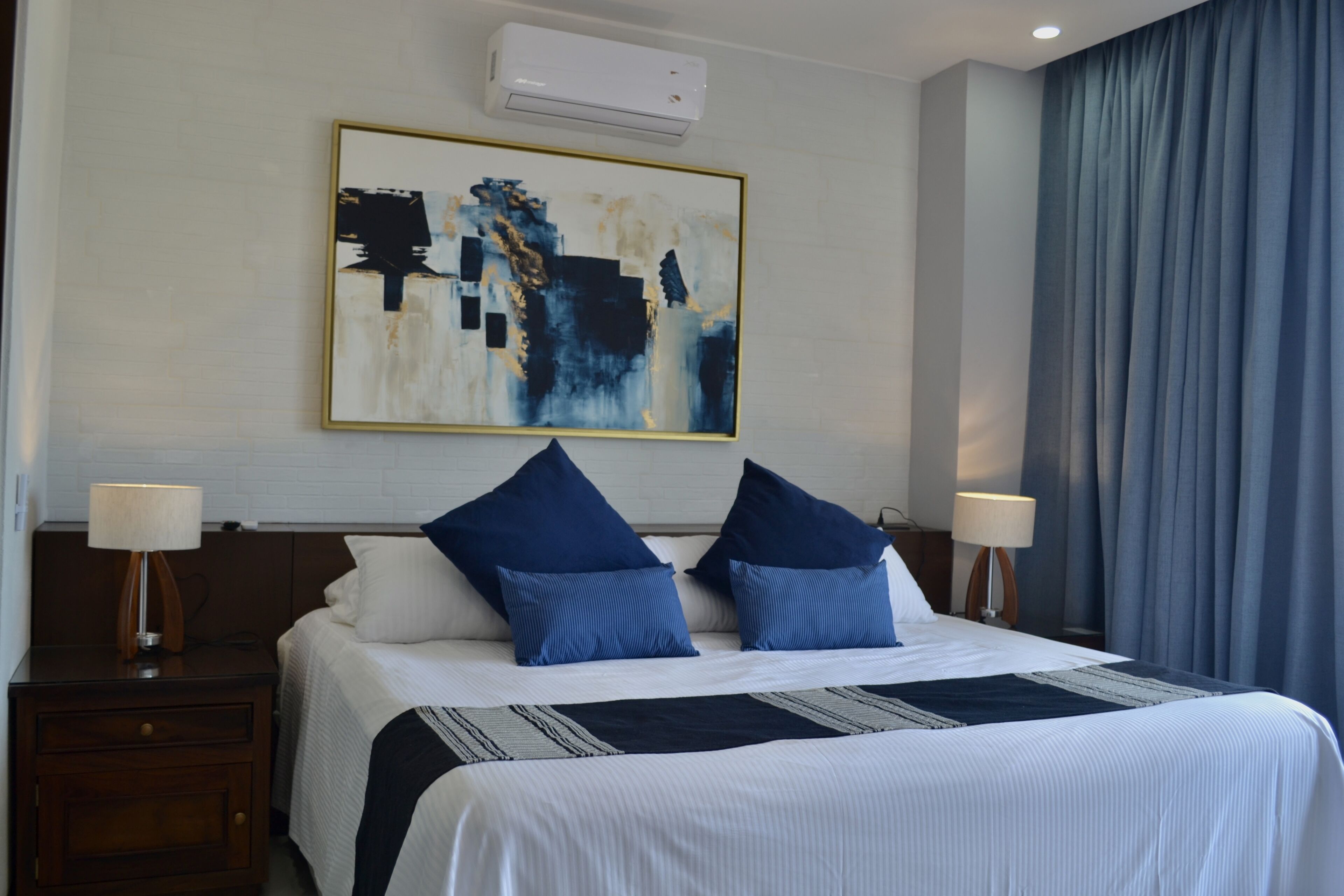 Habitación de hotel con una cama grande, dos mesitas de noche con lámparas, un aire acondicionado instalado en la pared y un cuadro abstracto de gran tamaño sobre la cama.