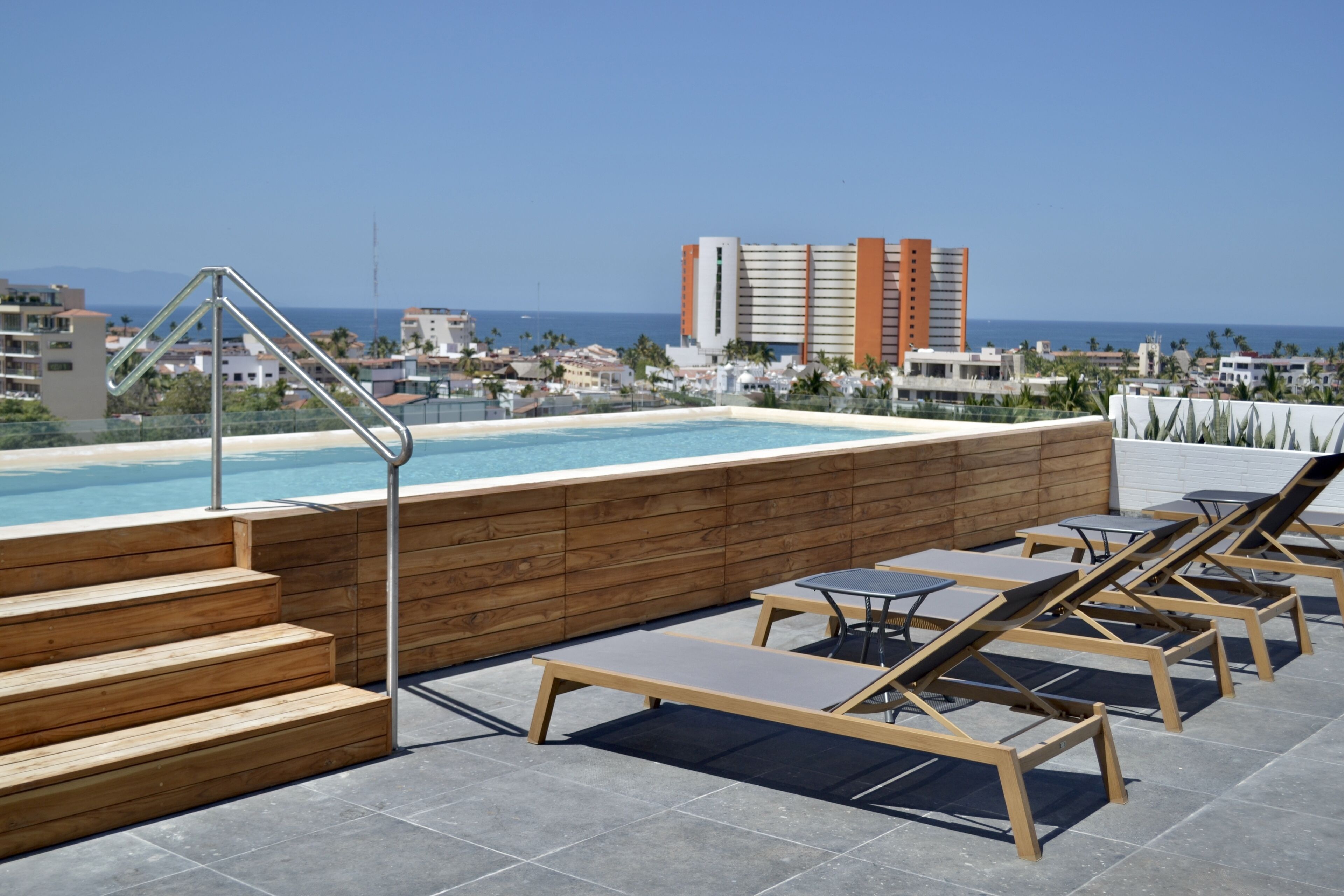 Ein Poolbereich auf dem Dach mit Holzstufen, Liegestühlen und Blick auf das Meer und Gebäude im Hintergrund.