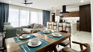 A modern dining area with a wooden table set for four, a kitchen in the background, and a balcony with curtains. - Amazing views coastal apartment. 2BD (Puerto Vallarta)