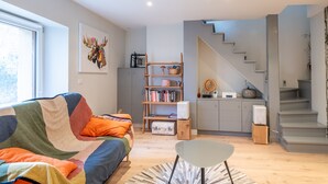 A modern living room with a colorful sofa, a glass coffee table, and a bookshelf. A staircase leads to a kitchen area.