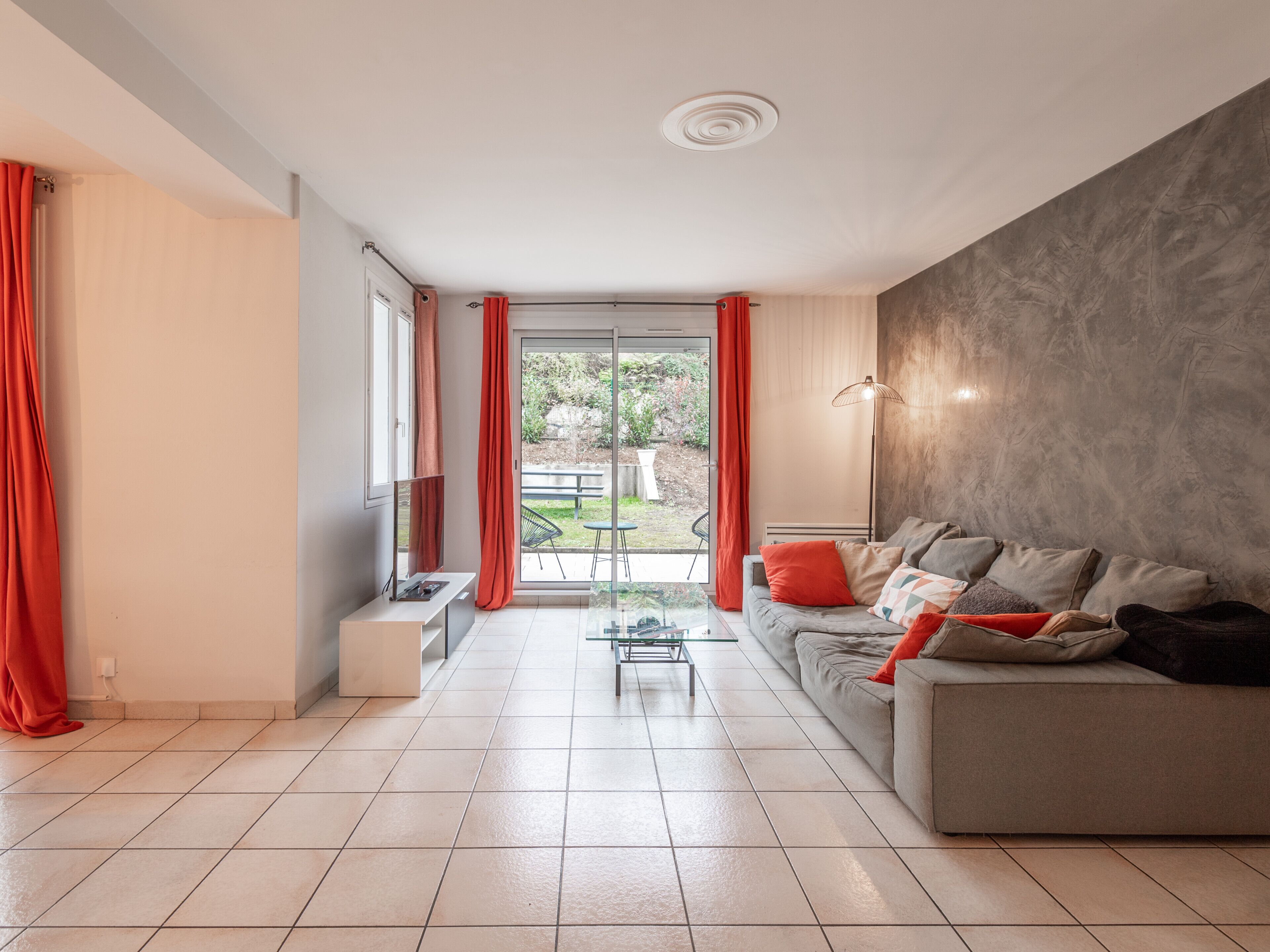 Een moderne woonkamer met een grijze bank, rode kussens, een glazen salontafel en uitzicht op een tuin via een schuifpui.