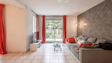 Een moderne woonkamer met een grijze bank, rode kussens, een glazen salontafel en uitzicht op een tuin via een schuifpui.