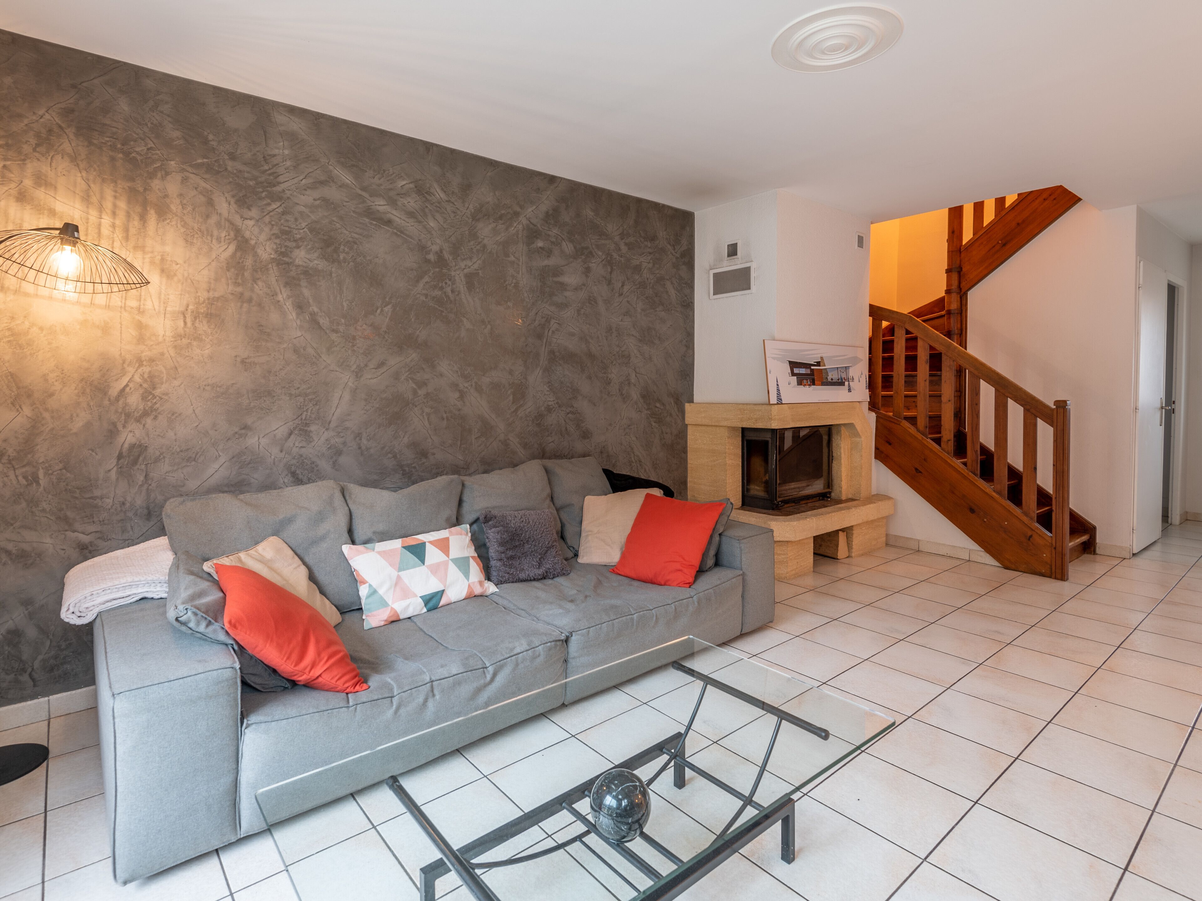 A living room with a grey sofa, a glass coffee table, a fireplace, and a wooden staircase.