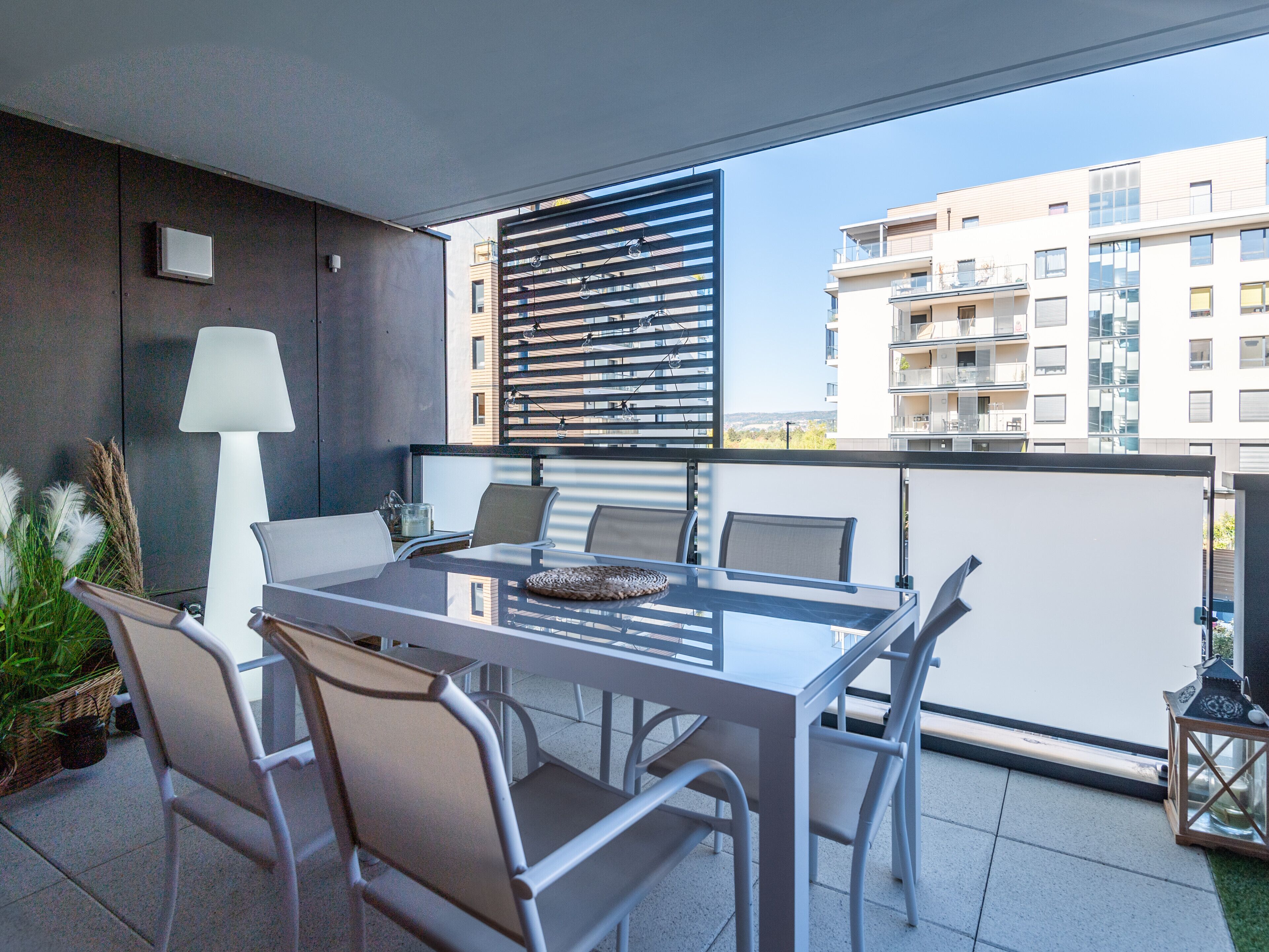 A balcony with a glass table and chairs, a white lamp, and a view of a modern building.