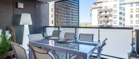 A balcony with a glass table and chairs, a white lamp, and a view of a modern building.