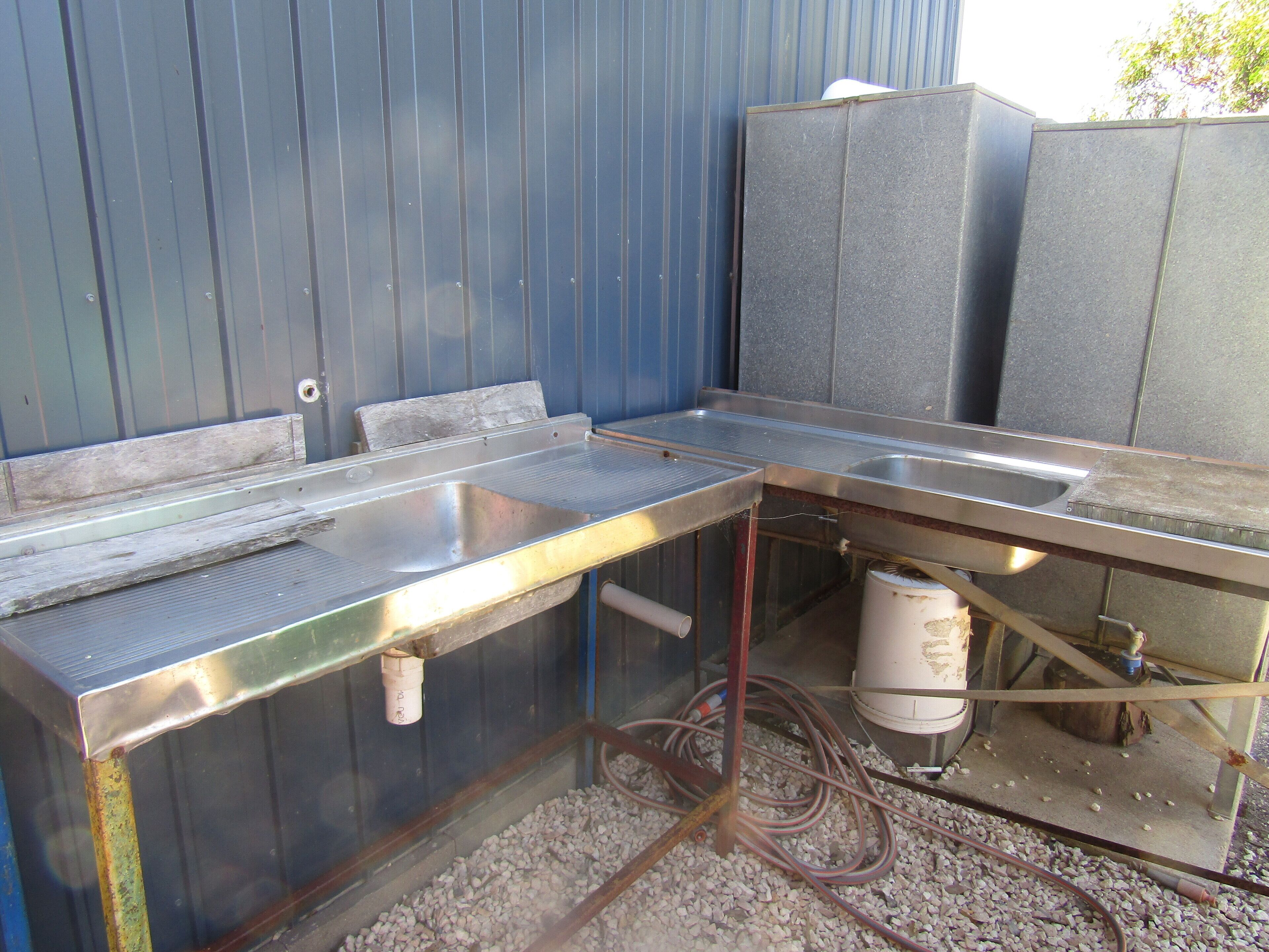 Cocina exterior de metal con dos lavabos, una manguera y un recipiente blanco.