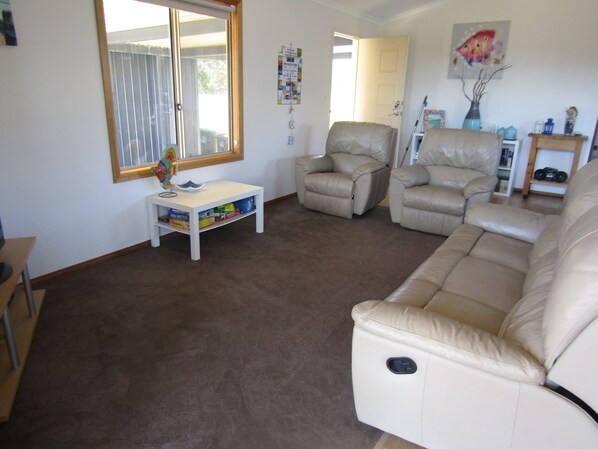A living room with a beige sofa, two recliner chairs, a small white table with toys, and a window with blinds.