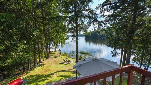 A deck with a grill and umbrella overlooking a lake surrounded by trees.
